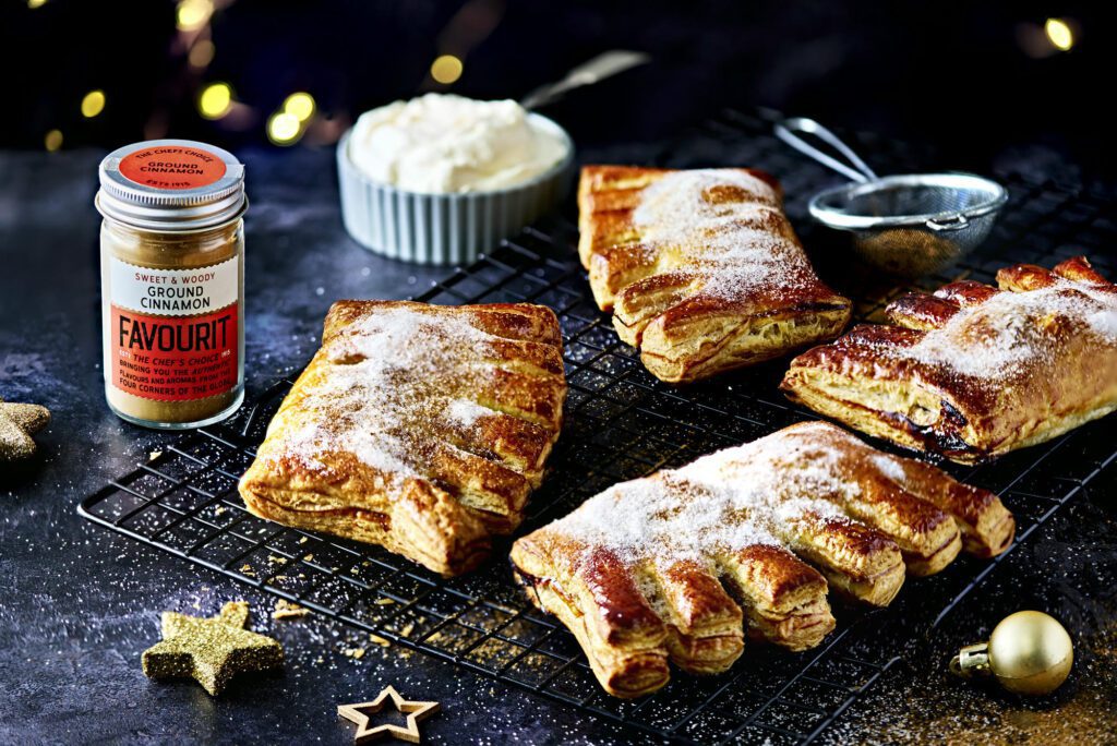 Five sugar-dusted bear claw pastries rest on a cooling rack, accompanied by a container of ground cinnamon, a bowl of cream, and decorative stars on the dark surface, evoking the warmth and charm of Christmas.