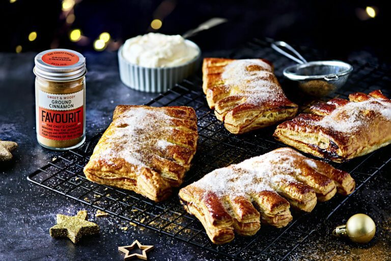 Five sugar-dusted bear claw pastries rest on a cooling rack, accompanied by a container of ground cinnamon, a bowl of cream, and decorative stars on the dark surface, evoking the warmth and charm of Christmas.