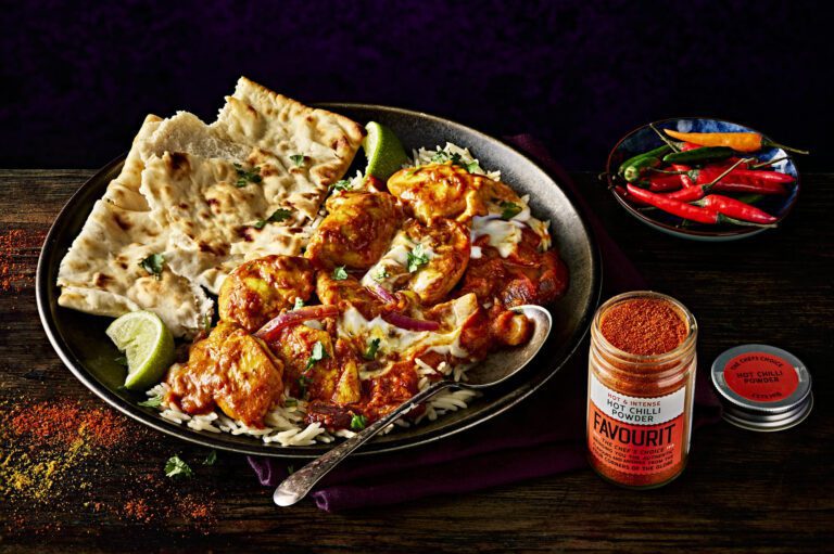A plate of spicy chicken curry sits alongside rice, naan, and lime wedges. Nearby, a bowl of red and green chilies and a jar of chili powder rest on the dark wooden table. This quick and flavorful dish offers a healthy twist reminiscent of traditional Chicken Tikka Masala.