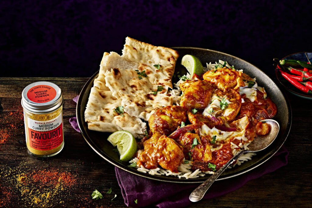 A plate featuring quick curry chicken with rice and naan bread, garnished with lime wedges and herbs. A jar of curry powder and red chilies rest nearby on a dark wooden surface, reminiscent of a vibrant Chicken Tikka Masala spread.