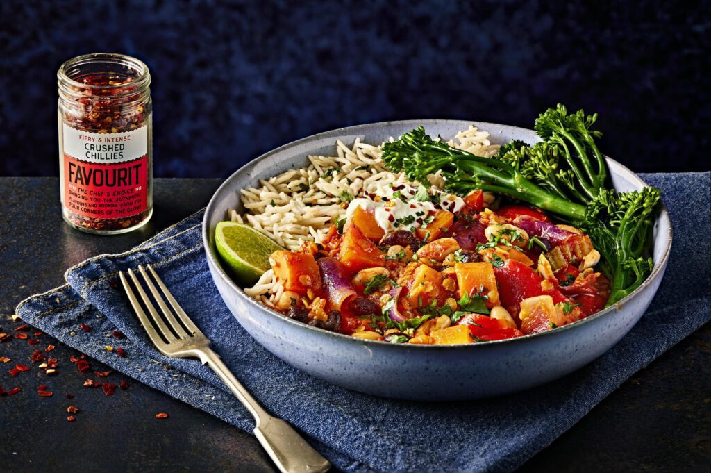 Bowl of hearty vegetable stew with rice and broccoli, garnished with lime and herbs, sits next to a jar of crushed chillies on a blue cloth. Lentils add an extra layer of richness to this nutritious delight.