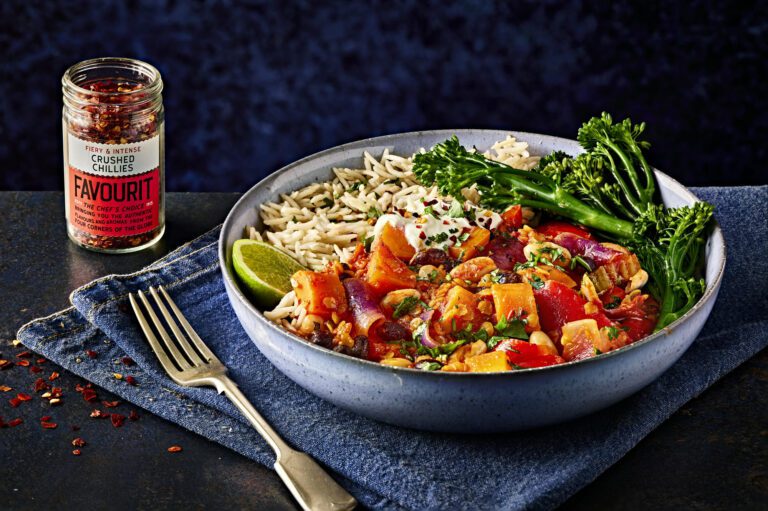 Bowl of hearty vegetable stew with rice and broccoli, garnished with lime and herbs, sits next to a jar of crushed chillies on a blue cloth. Lentils add an extra layer of richness to this nutritious delight.