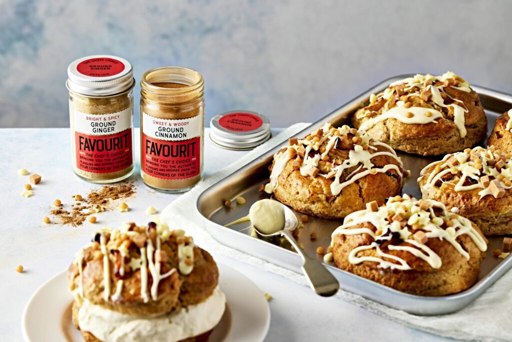 A tray of buttermilk scones with icing and chopped nuts sits next to jars of ground ginger and cinnamon. A spiced apple scone adorned with cream and nuts steals the spotlight on a plate in the foreground.