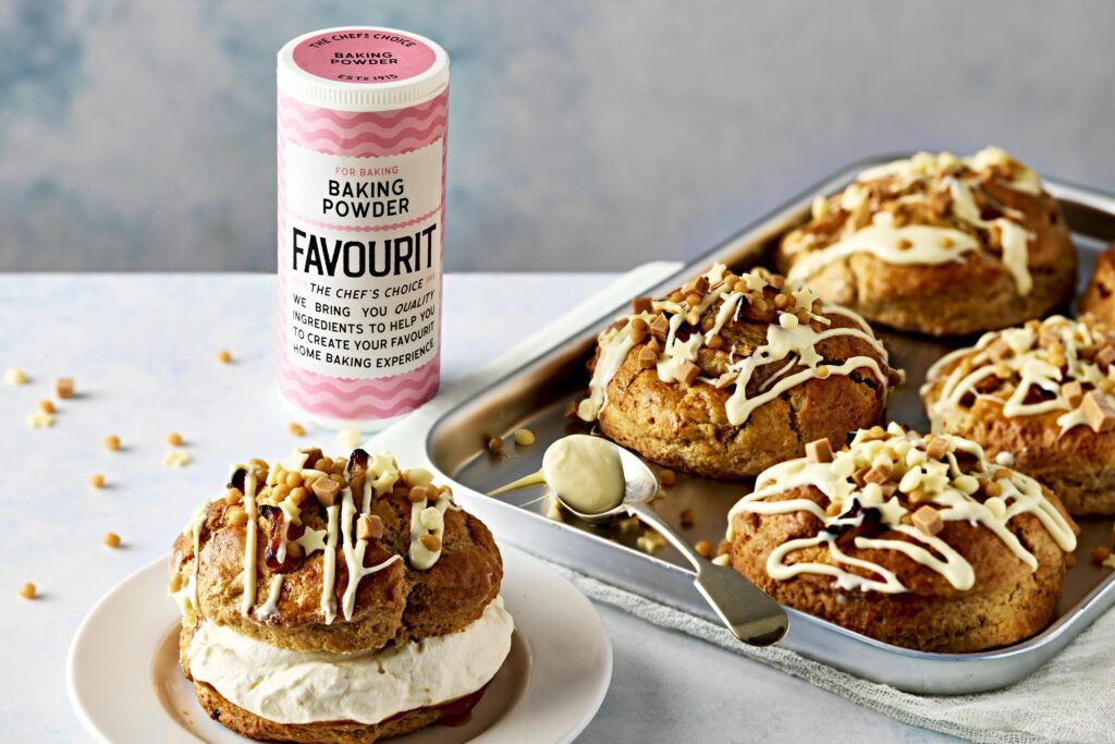A baking powder container sits beside a tray of delightful buttermilk scones. These scones are topped with white icing and nuts, while one is filled with cream.