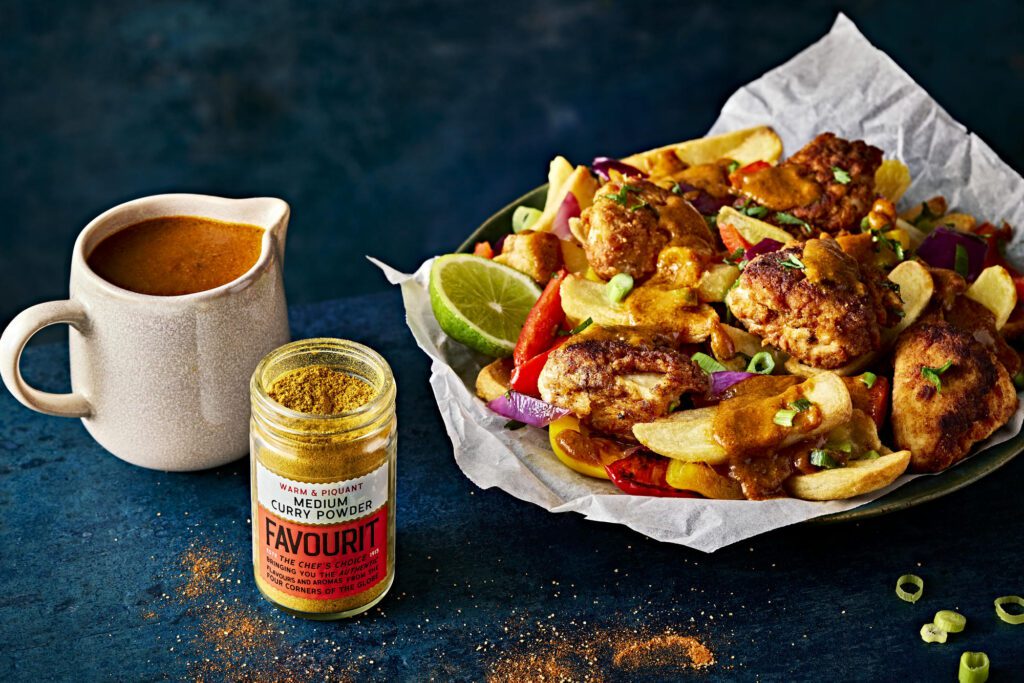 A plate of Salt & Chilli Chicken wings with sliced vegetables and sauce, accompanied by a jug of sauce and a jar labeled "curry powder" sit enticingly on a dark surface, ready to be devoured.