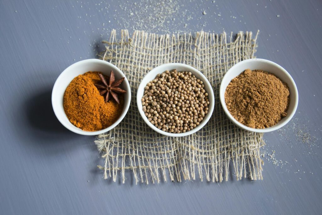 Three white bowls on a woven mat: one with ground turmeric and star anise, one with coriander seeds, and one with ground cumin. This display is a testament to the art of spice blending, expertly curated for those who value a professional touch in their culinary endeavors.