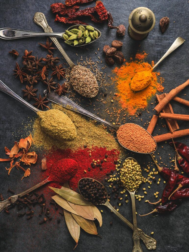 An assortment of spices and herbs on a dark surface with spoons, including cardamom, turmeric, cumin, coriander, bay leaves, and chili peppers.