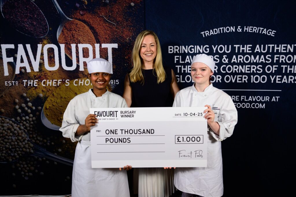Three people pose holding a large cheque for one thousand pounds at the FAVOURIT anniversary event; two young chefs in uniform flank a smiling woman in the center, all looking proudly at the camera.