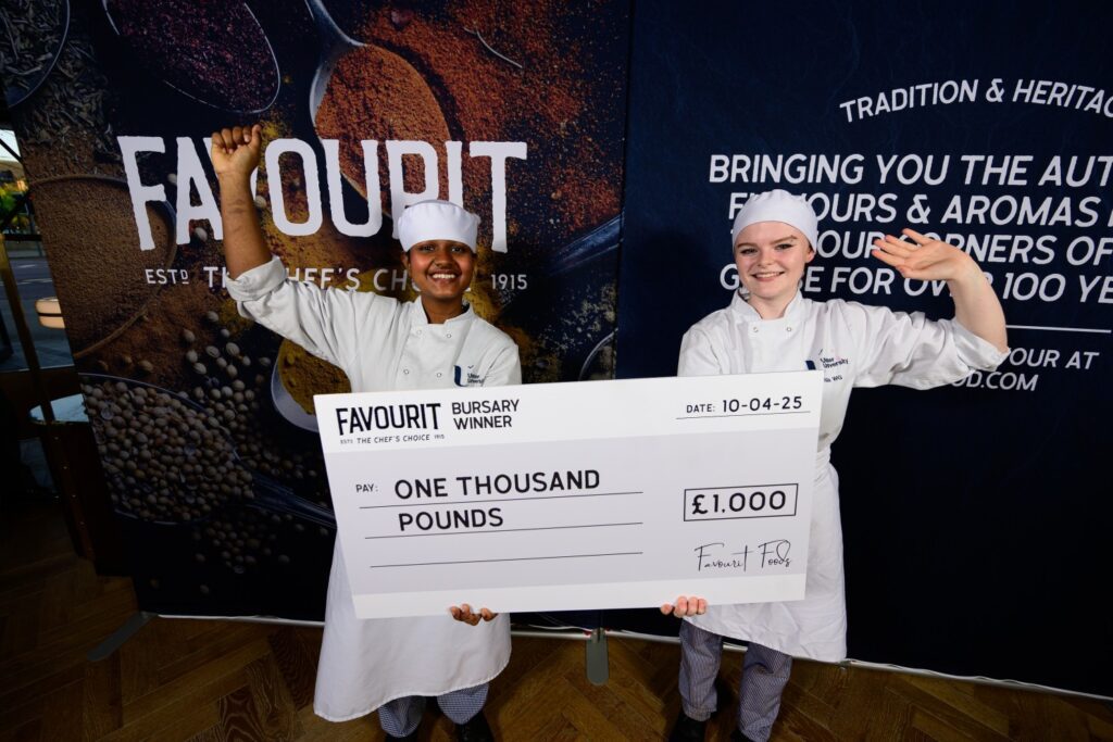 Two young chefs in uniform stand smiling and waving, holding a large check for one thousand pounds awarded by "Favourit" at an anniversary event dated 10-04-25.