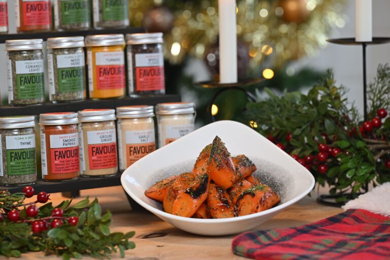 A white bowl of roasted carrots garnished with herbs sits on a wooden table beside a rack of spice jars, festive greenery, and a red plaid napkin.