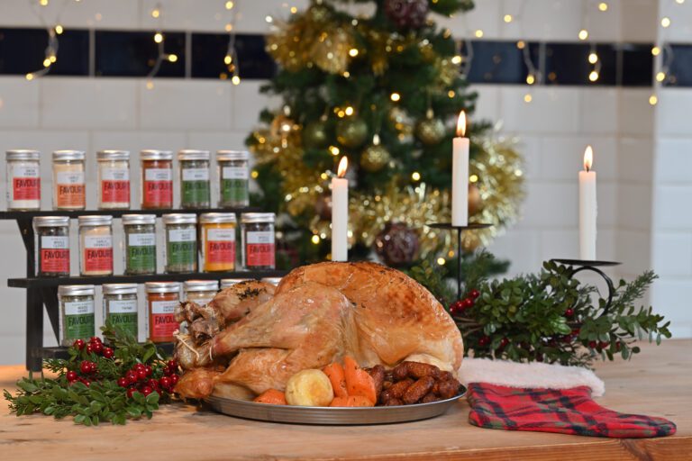 A roast turkey with vegetables on a platter sits on a wooden table, surrounded by candles, spices, a plaid napkin, and holiday greenery, with a decorated Christmas tree in the background.
