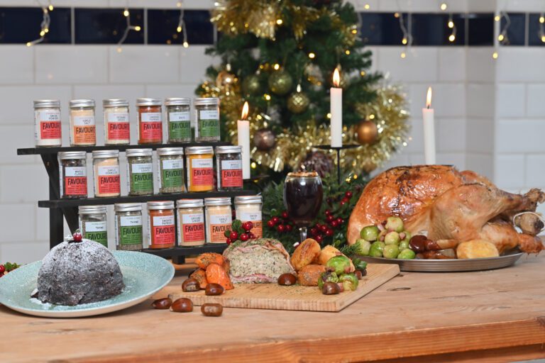 A table set with a roast turkey, vegetables, a Christmas pudding, assorted spices in jars, and a decorated Christmas tree in the background.