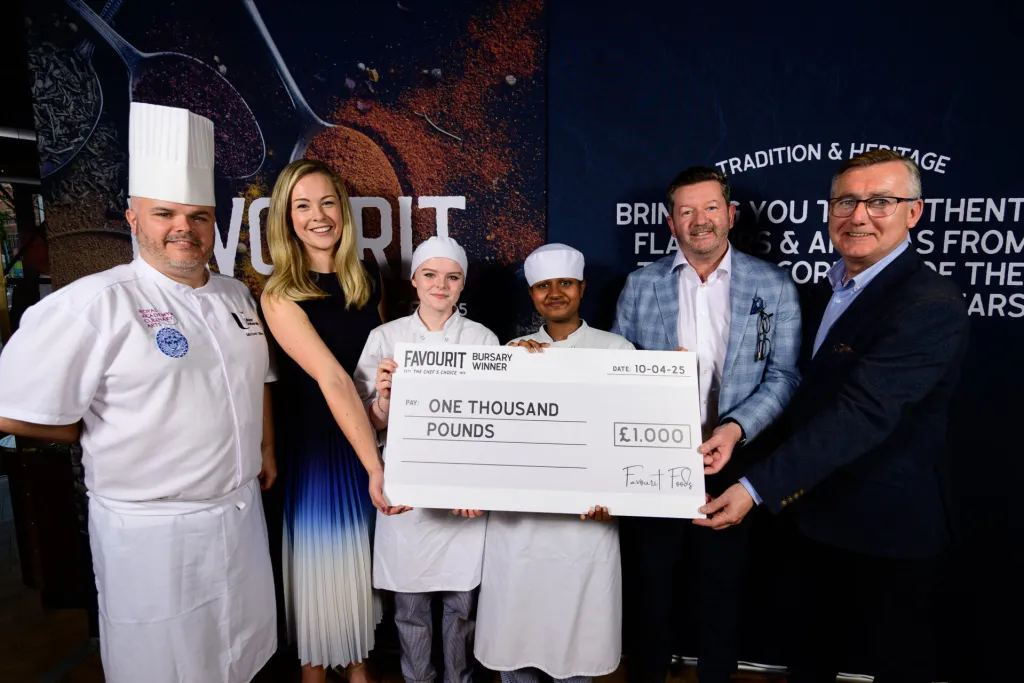 Five people, including two young chefs in uniform, hold a large check for £1,000 labeled "FAVOURIT Bursary Winner" while posing for a photo at the anniversary event.