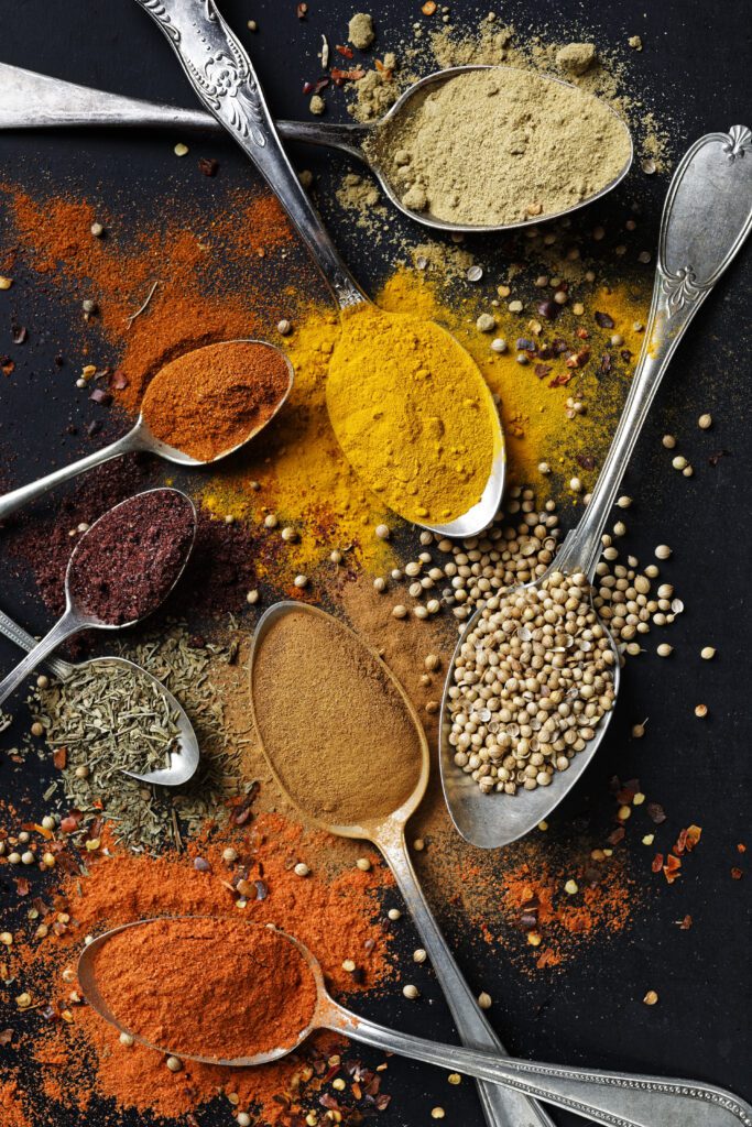 5 metal spoons laid out on a table with various different herbs and spices on them. They are vibrant colours like yellow, orange, and green.